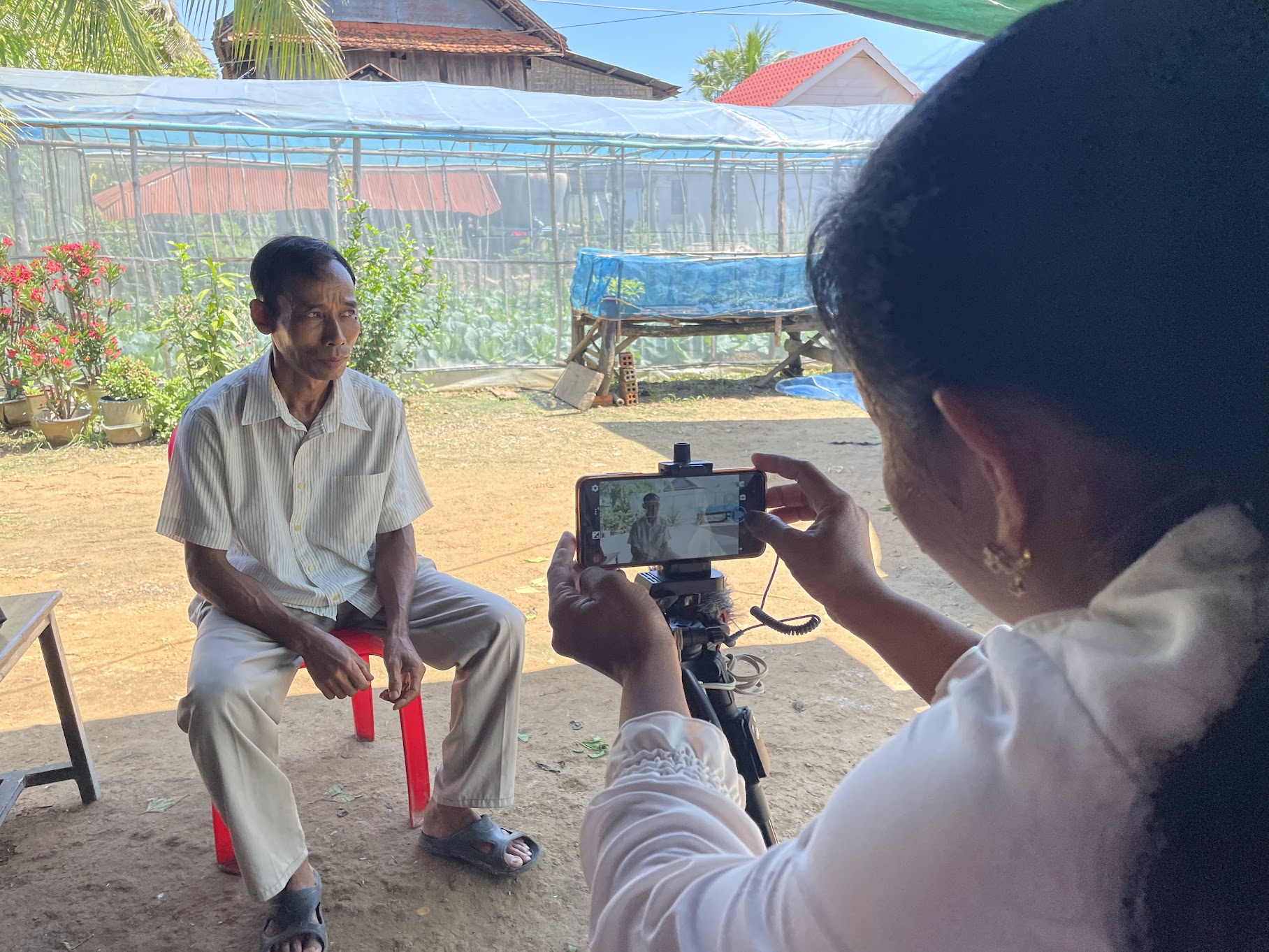 A woman recording a video on a smartphone. A farmer is seated on a chair in front of her